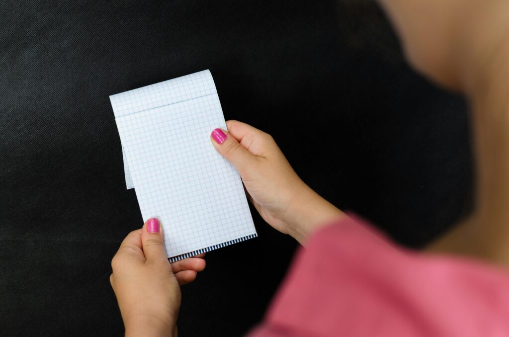 A close-up of a woman holding a small notepad with checked paper, against a dark background.